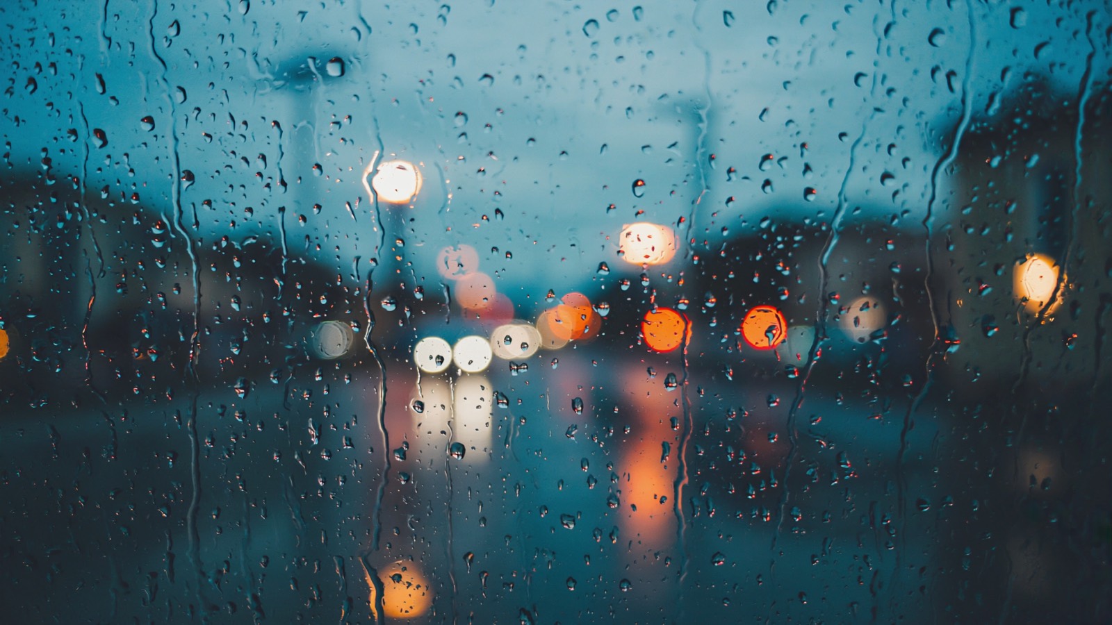 Regennasse Fensterscheibe von innen, dahinter unscharfe warme Lichter einer fremden Stadt bei Dämmerung
