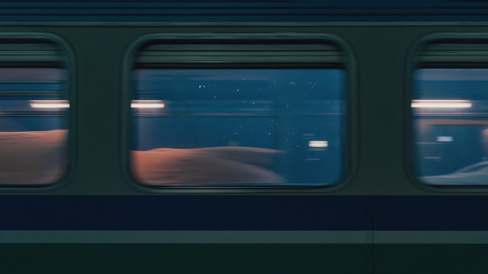 Blick aus einem dunklen Zugabteilfenster in die Nacht, verschwommene Lichter ferner Häuser ziehen als Streifen vorbei